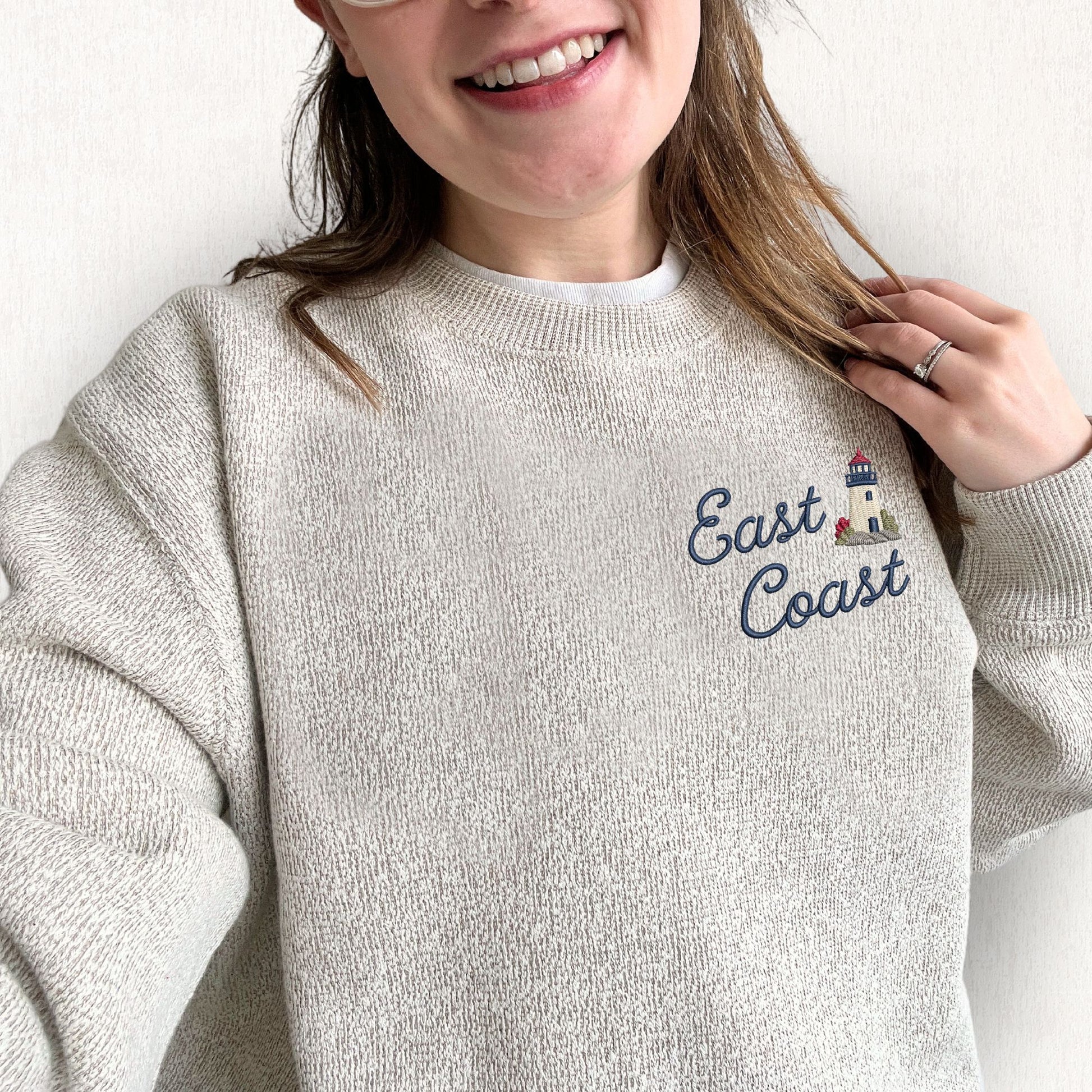 A woman wearing an oatmeal heather knit crewneck sweatshirt with 'East Coast' embroidered in navy script next to a small lighthouse graphic on the left chest.