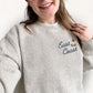 A woman wearing an oatmeal heather knit crewneck sweatshirt with 'East Coast' embroidered in navy script next to a small lighthouse graphic on the left chest.