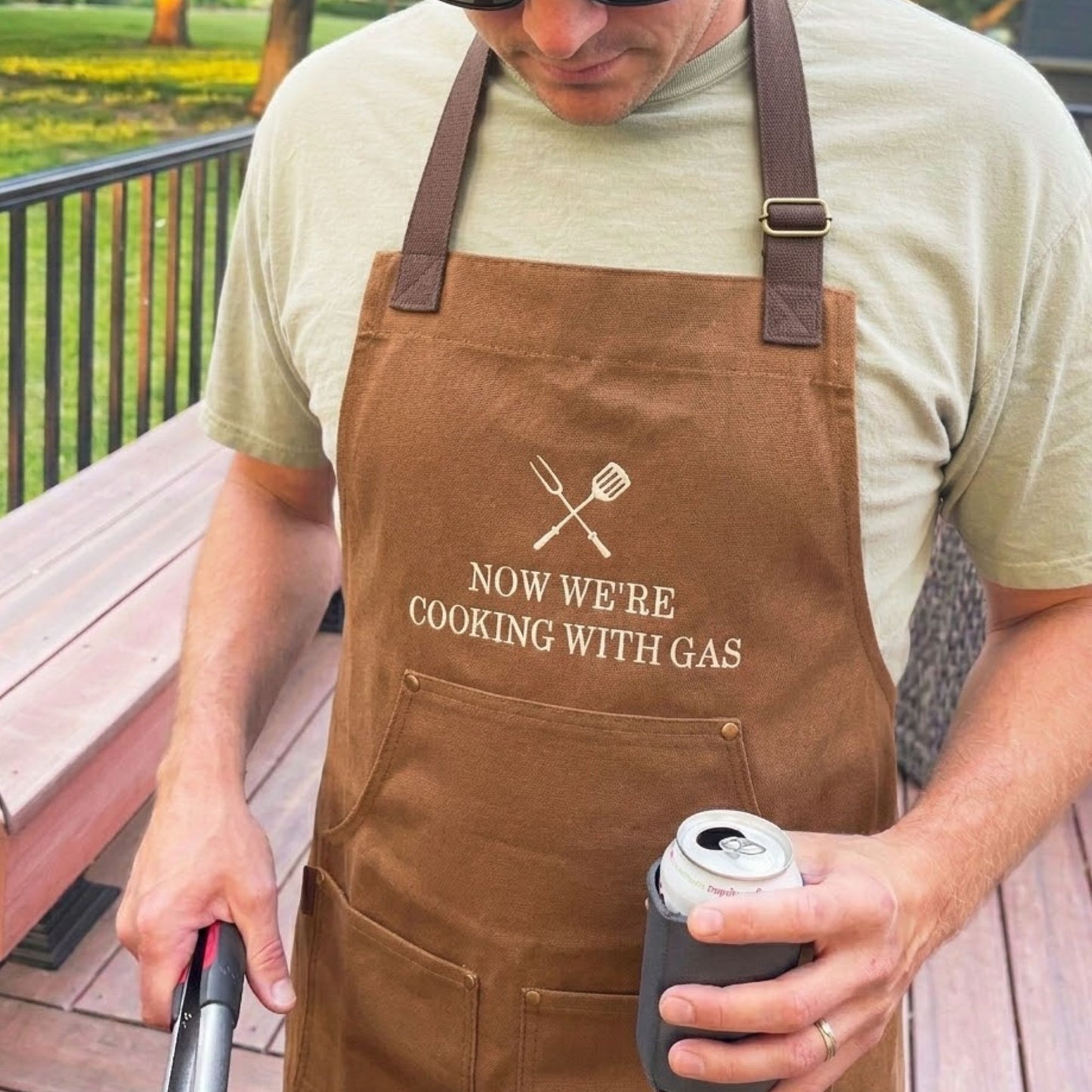 Man wearing a brown apron with text, grilling and holding a can on a wooden deck.