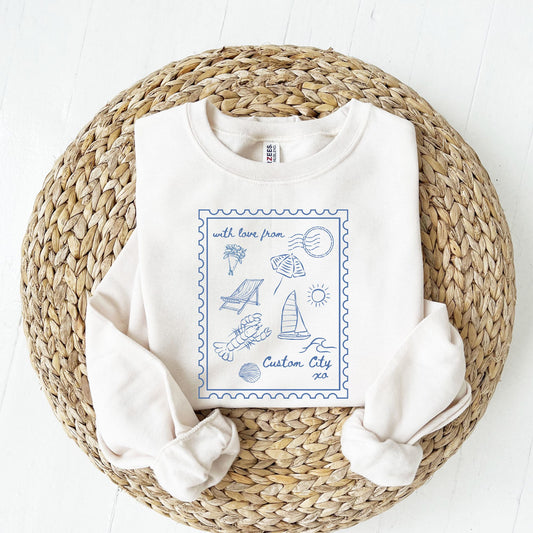 folded white sweatshirt with  a printed design featuring a stamp outline with lobster, sailboat, folding chair, clam, waves, bouquet of flowers, umbrella, XO, and a space to add a custom city.