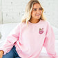 young woman wearing a light pink crewneck sweatshirt with embroidered  pink mug with marshmallows a ginger breadman and candy cane on the left chest.