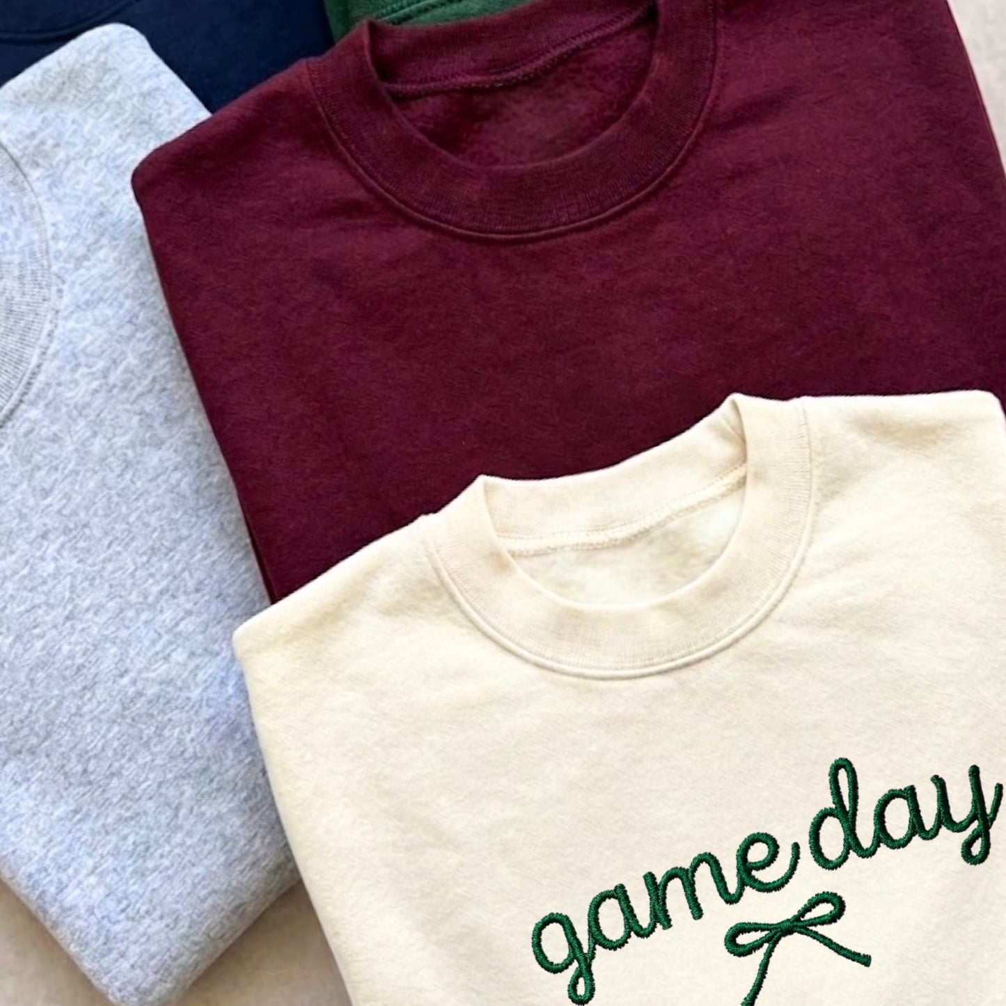 Three folded crewneck sweatshirts stacked diagonally: a light gray heather shirt on the left, a burgundy shirt in the back, and a cream shirt in the front with 'game day' embroidered in green script with a small bow.