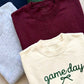 Three folded crewneck sweatshirts stacked diagonally: a light gray heather shirt on the left, a burgundy shirt in the back, and a cream shirt in the front with 'game day' embroidered in green script with a small bow.