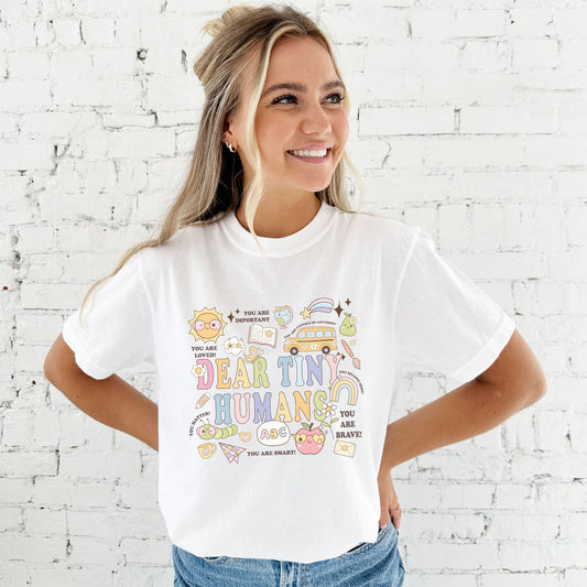 woman wearing a medium white comfort colors t-shirt with a colorful fun school icon dear tiny humans printed design on the front