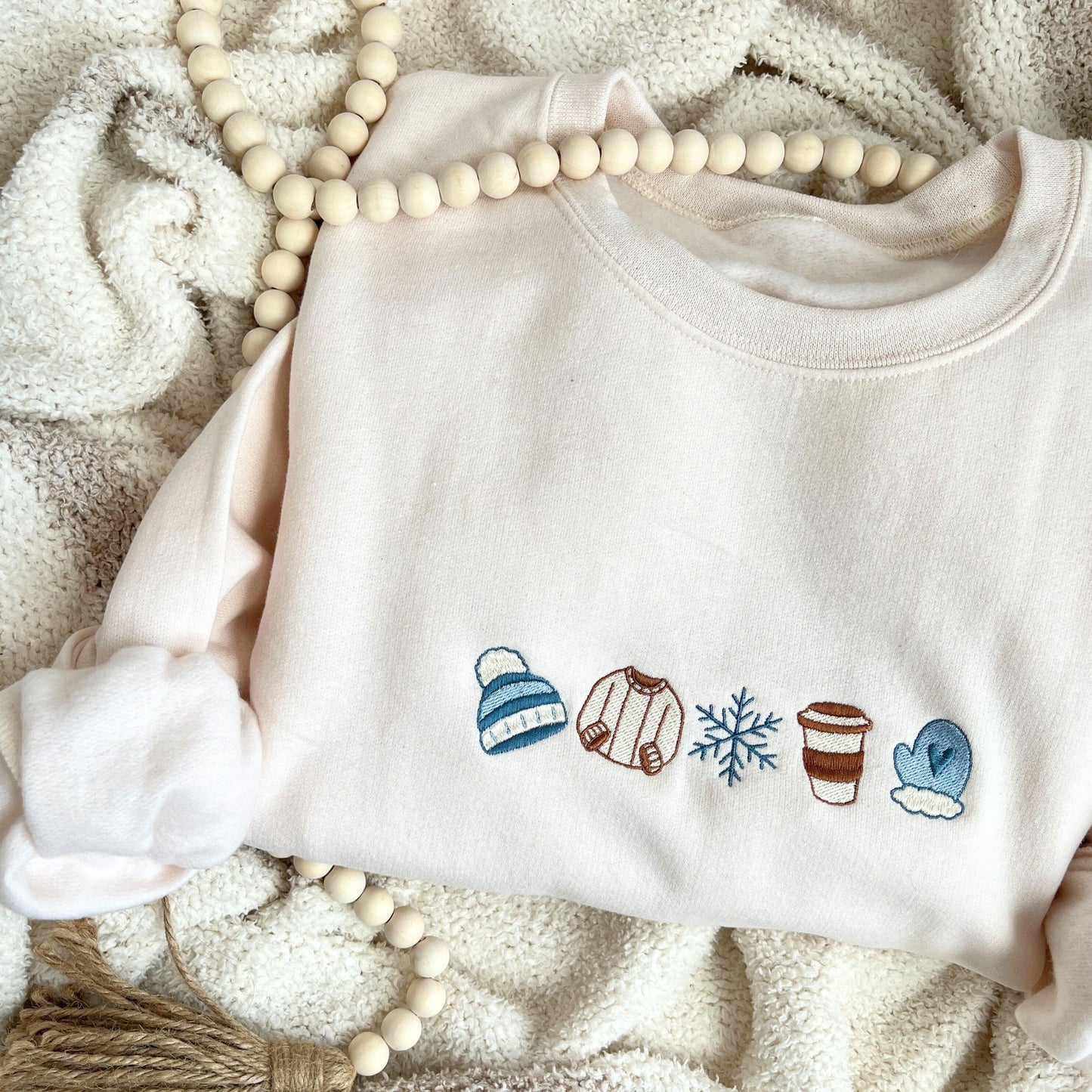 sweet cream crewneck sweatshirt with custom winter themed icons embroidered on the center chest. design featuring a cute beanie, sweater, snowflake, to-go coffee cup, and mitten in shades of blue, brown, and natural