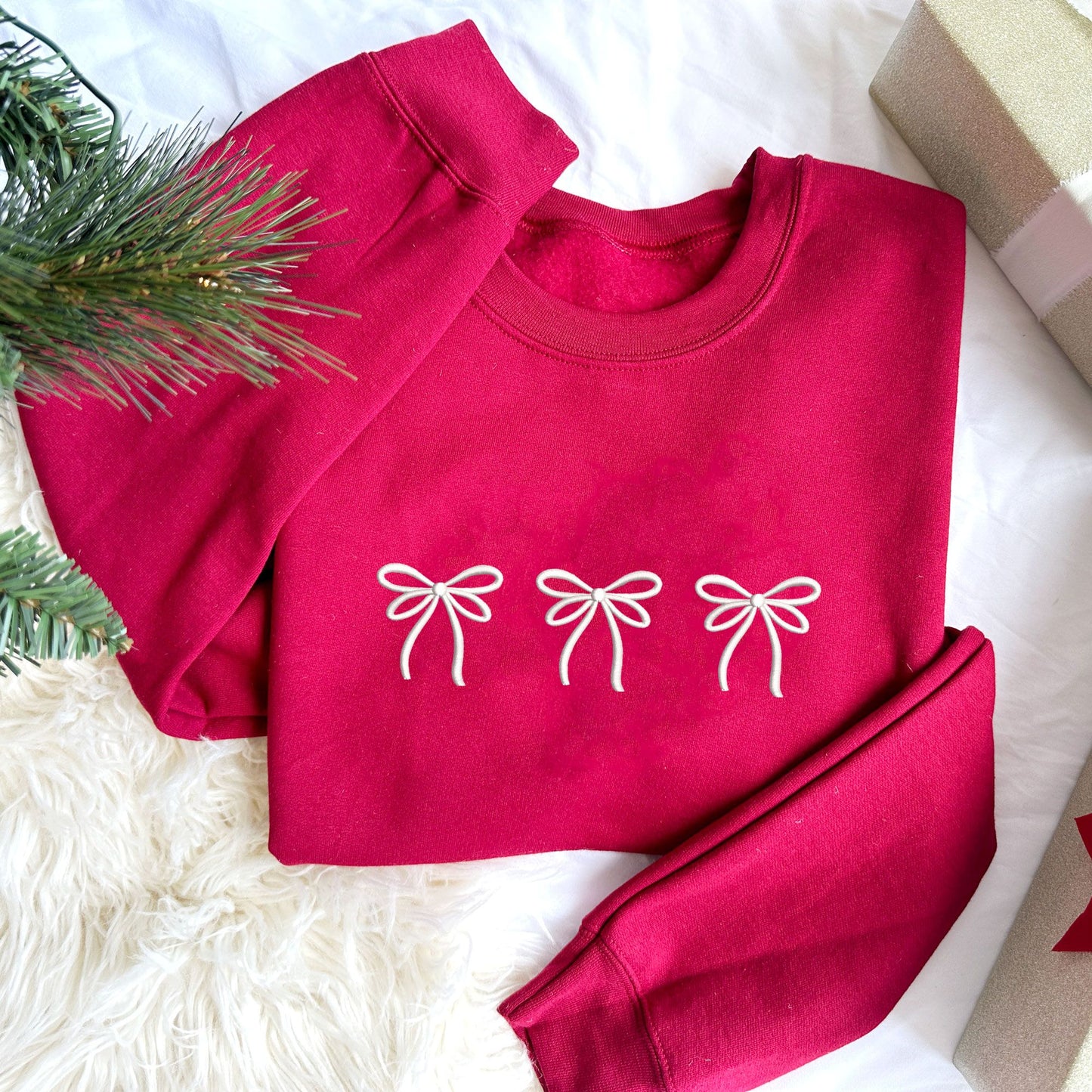 Christmas styled flat lay photo of a cardinal red crewneck with three embroidered bows across the chest in white thread.