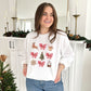 woman standing in front of a fireplace wearing a white pullover with a retro reindeer, bow, cocoa, tree, cookie, and snow globe printed design