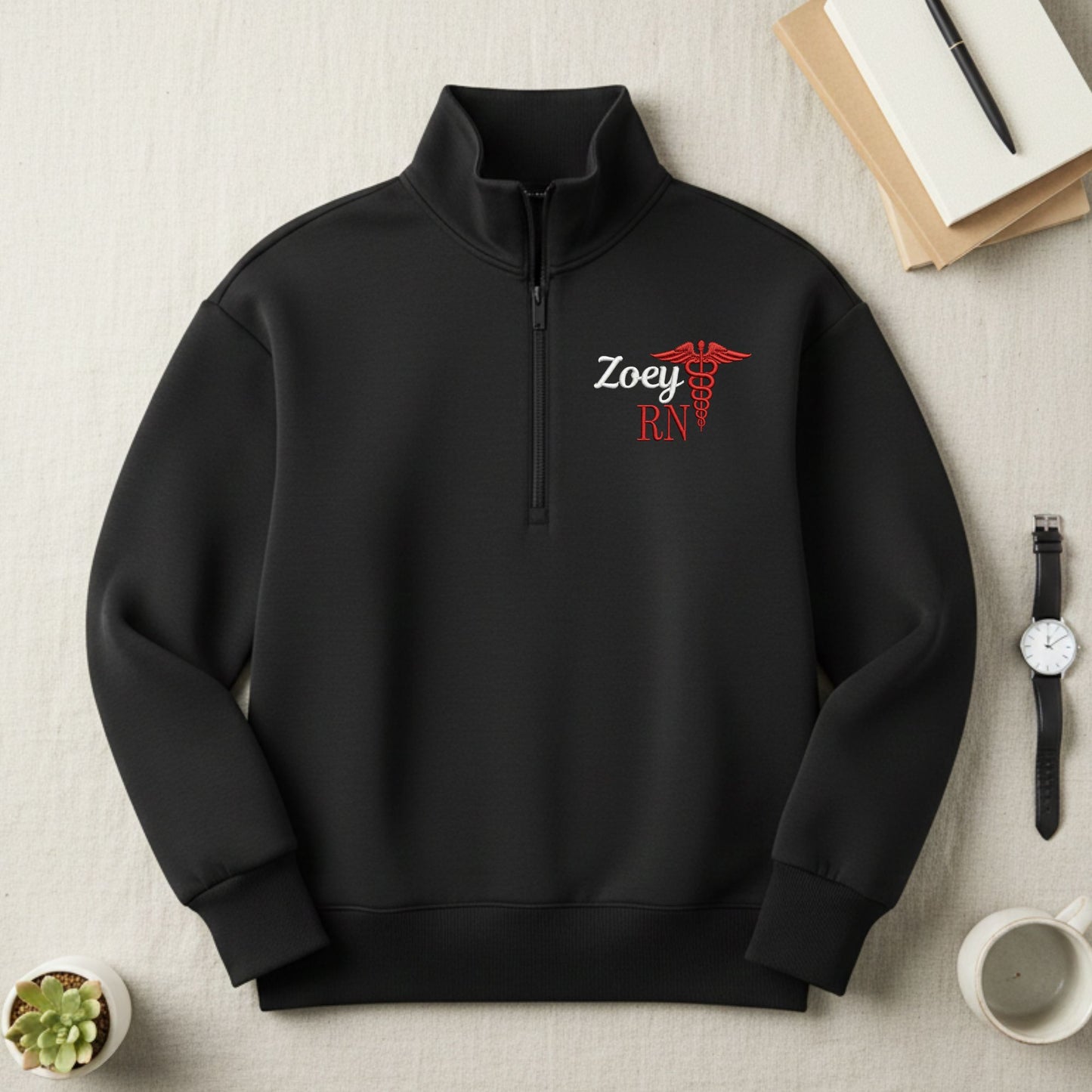 Black quarter zip sweatshirt with 'Zoey RN' design embroidered on the left chest on a light surface with books and a watch.