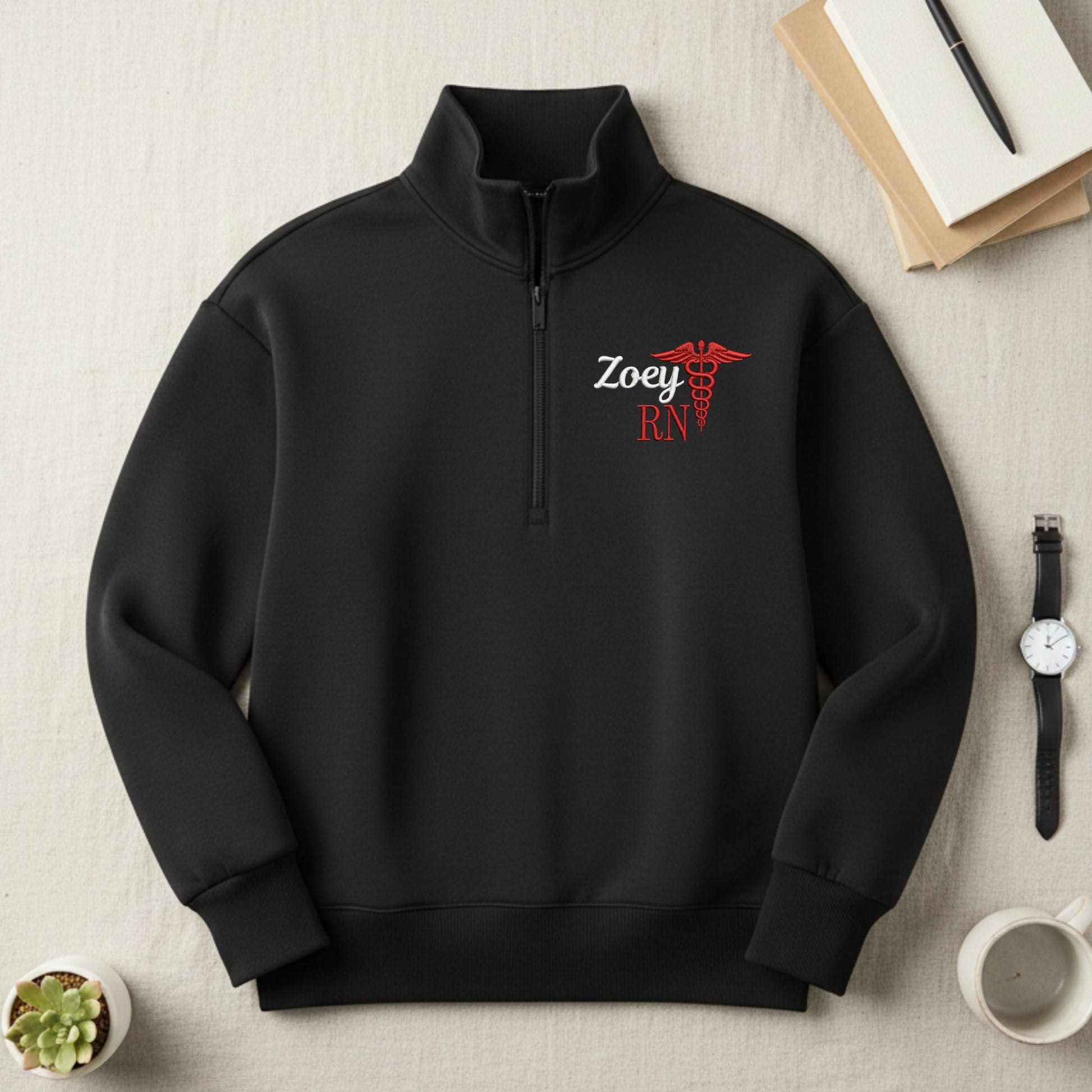 Black quarter zip sweatshirt with 'Zoey RN' design embroidered on the left chest on a light surface with books and a watch.