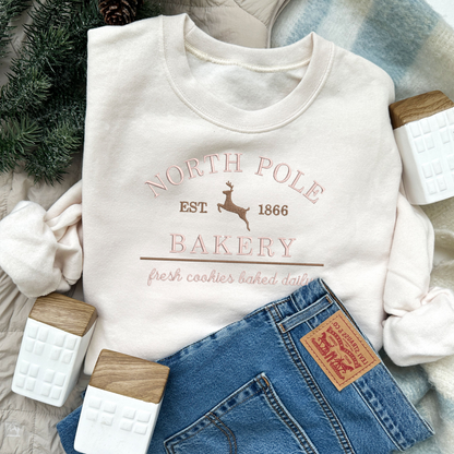 christmas styled flat lay of a sweet cream crewneck sweatshirt with embroidered north pole bakery design in pink and brown threads.