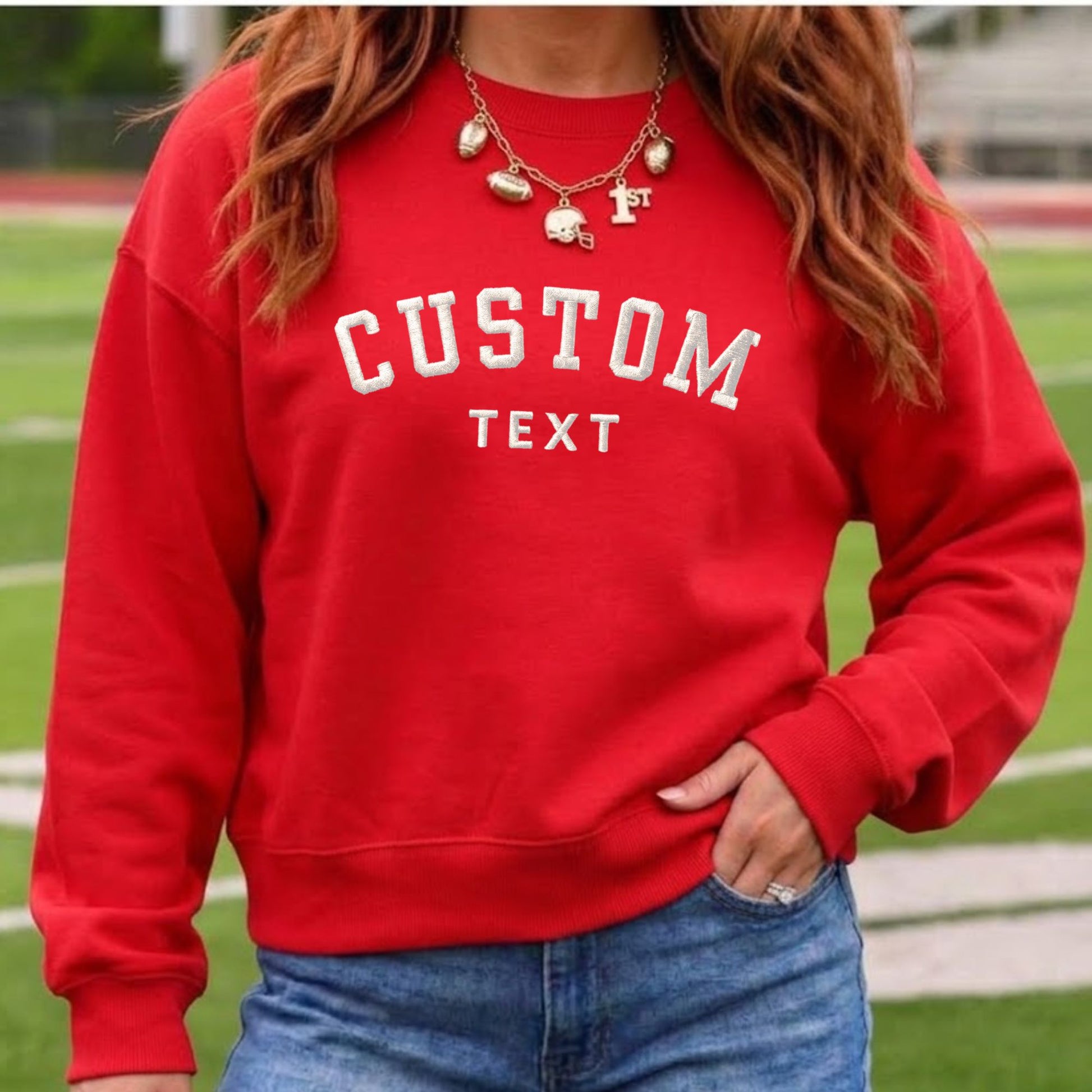 A woman wearing a bright red crewneck sweatshirt with 'CUSTOM TEXT' embroidered in white varsity-style letters on the front, paired with blue jeans and layered gold necklaces, set against a blurred green athletic field background.