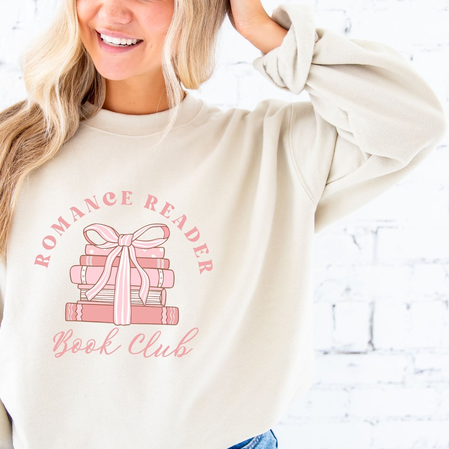 woman wearing a sand crewneck sweatshirt with a romance reader book club pink bow printed design