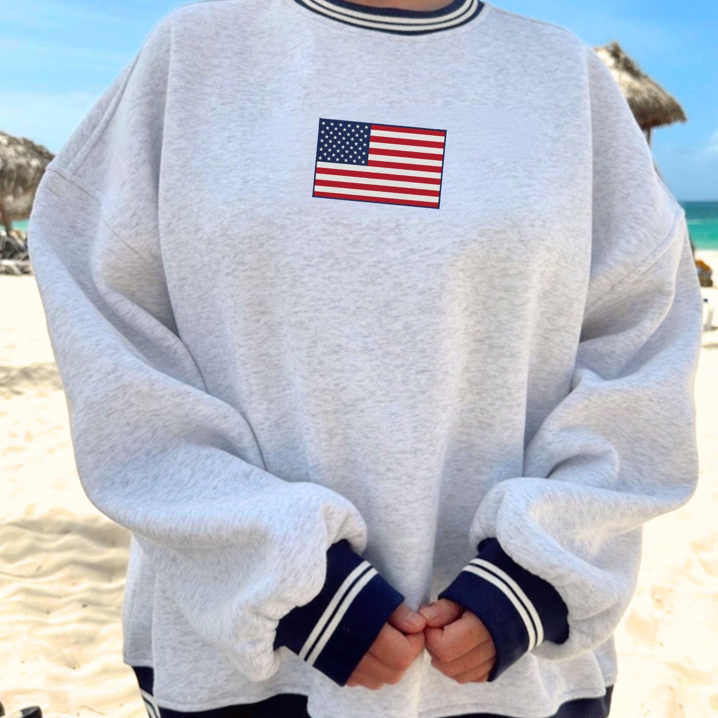 Person wearing a gray sweatshirt with an American flag embroidery on a beach.