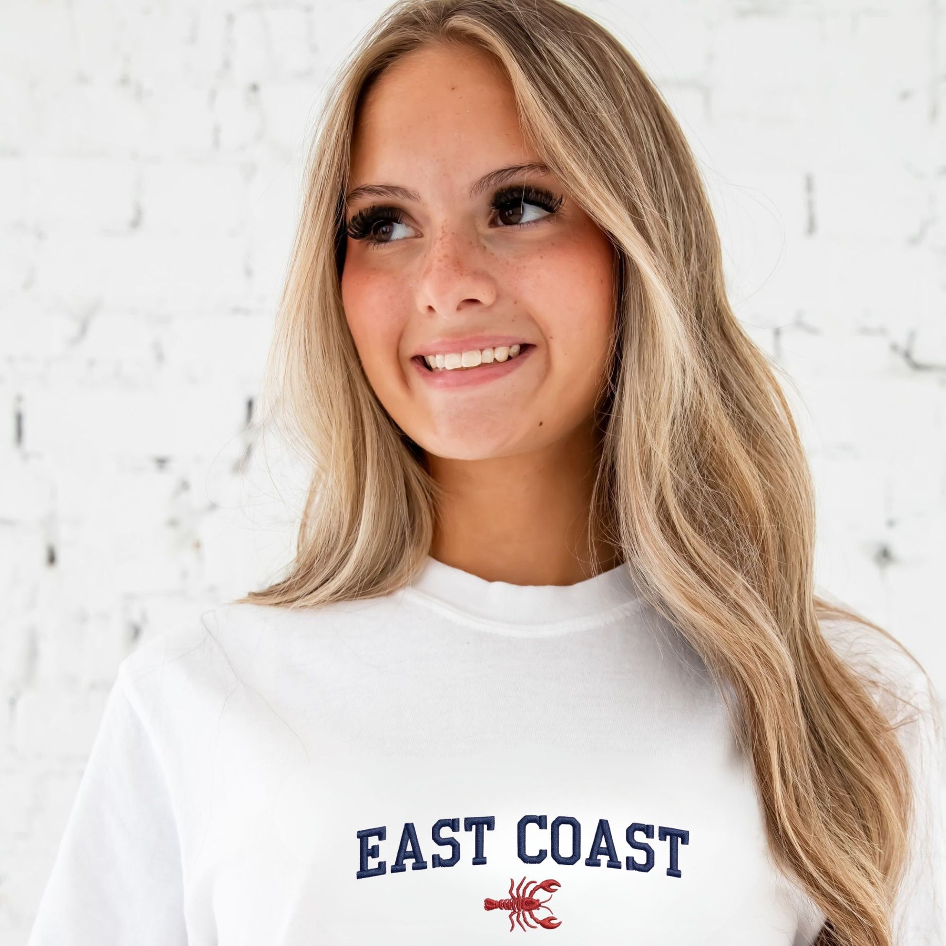 Woman wearing a white sweatshirt with 'East Coast' and a lobster graphic on a light background