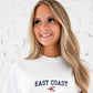 Woman wearing a white sweatshirt with 'East Coast' and a lobster graphic on a light background