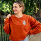 woman wearing a texas orange crewneck sweatshirt with embroidered ghost drinking iced coffee and holding a jack-o-lantern on the left chest