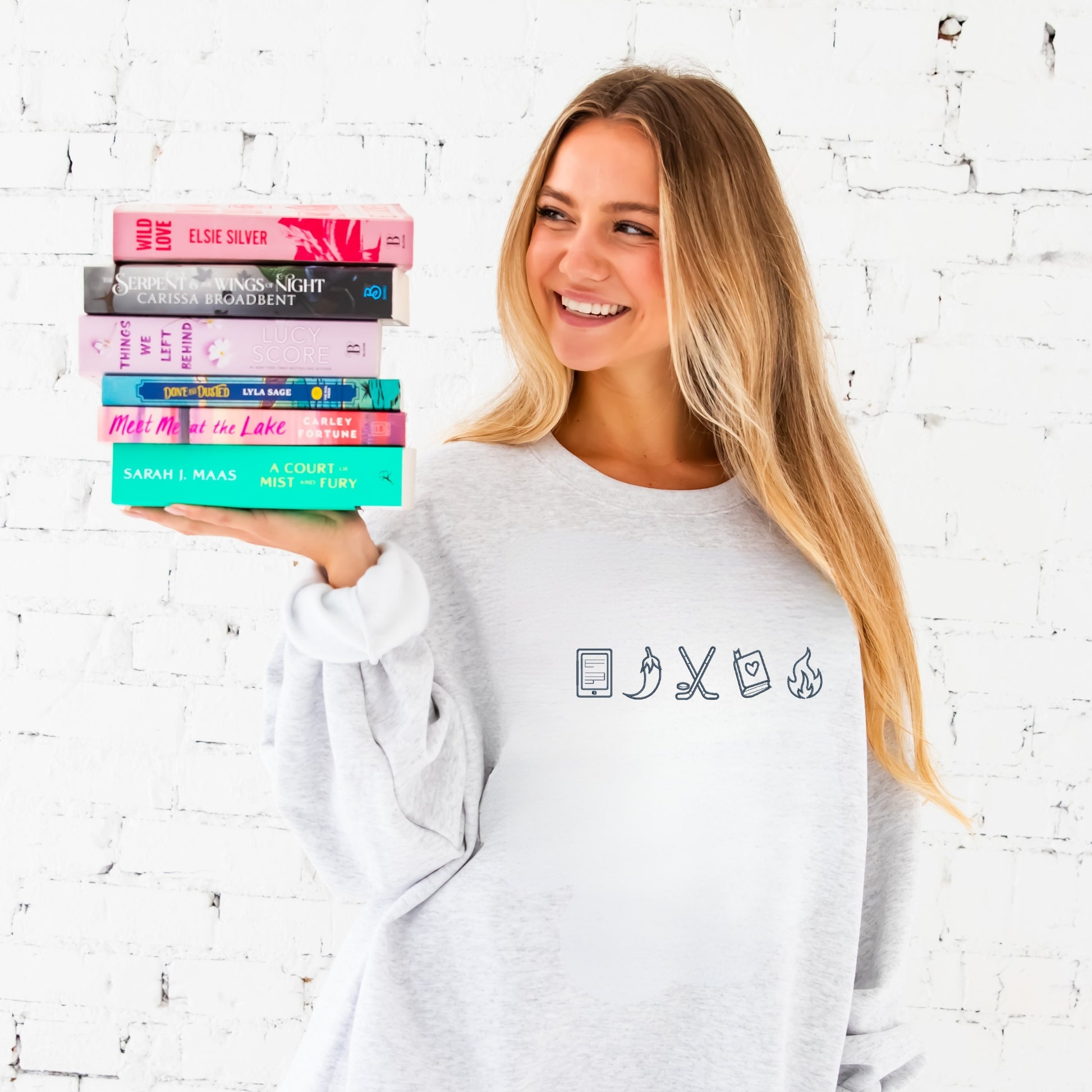 Woman holding a stack of books against a white brick wall. shes wearing an ash crewneck with a kindle, spicy pepper, hockey sticks, book, and fire icon design embroidered on the center chest