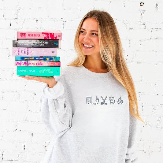 Woman holding a stack of books against a white brick wall. shes wearing an ash crewneck with a kindle, spicy pepper, hockey sticks, book, and fire icon design embroidered on the center chest