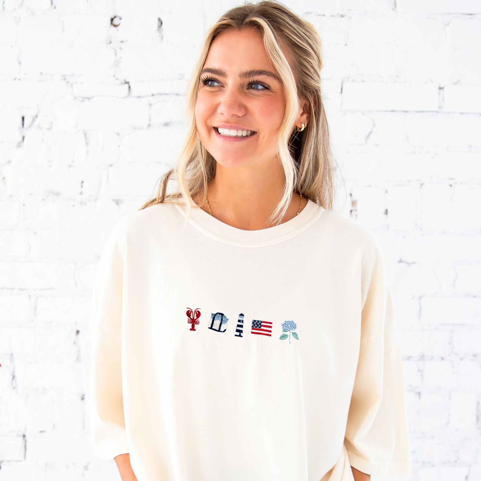 Woman wearing a cream-colored sweatshirt with embroidered icons against a white brick wall.
