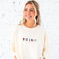 Woman wearing a cream-colored sweatshirt with embroidered icons against a white brick wall.