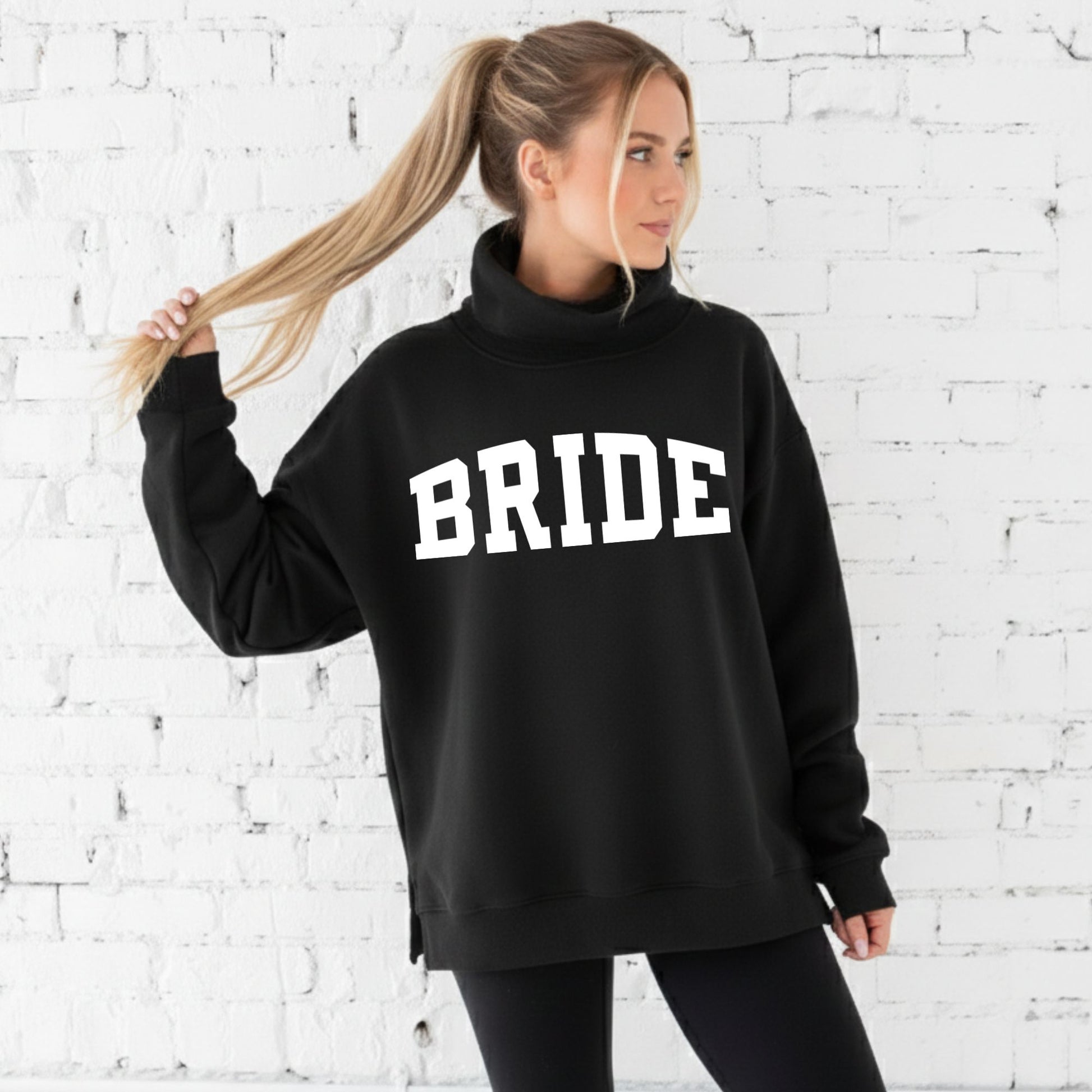 Woman wearing an oversized black mockneck sweatshirt with 'BRIDE' text against a white brick wall.