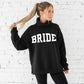 Woman wearing an oversized black mockneck sweatshirt with 'BRIDE' text against a white brick wall.