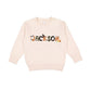 Natural heather crewneck sweatshirt with a custom name print in a mixed patterned Halloween text and accented with cute spooky icons like bats, a ghost, jack-o-lantern, and witch hat.