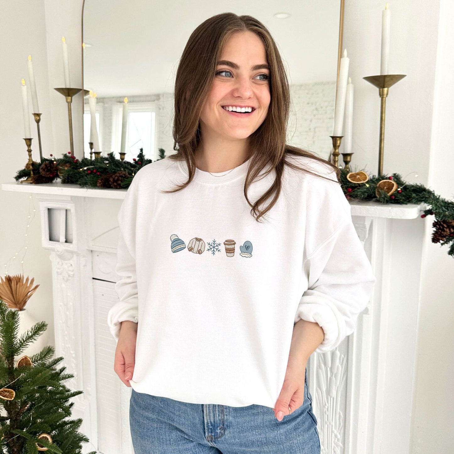 young woman wearing a white crewneck sweatshirt with custom winter themed icons embroidered on the center chest. design featuring a cute beanie, sweater, snowflake, to-go coffee cup, and mitten in shades of blue, brown, and natural