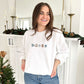 young woman wearing a white crewneck sweatshirt with custom winter themed icons embroidered on the center chest. design featuring a cute beanie, sweater, snowflake, to-go coffee cup, and mitten in shades of blue, brown, and natural