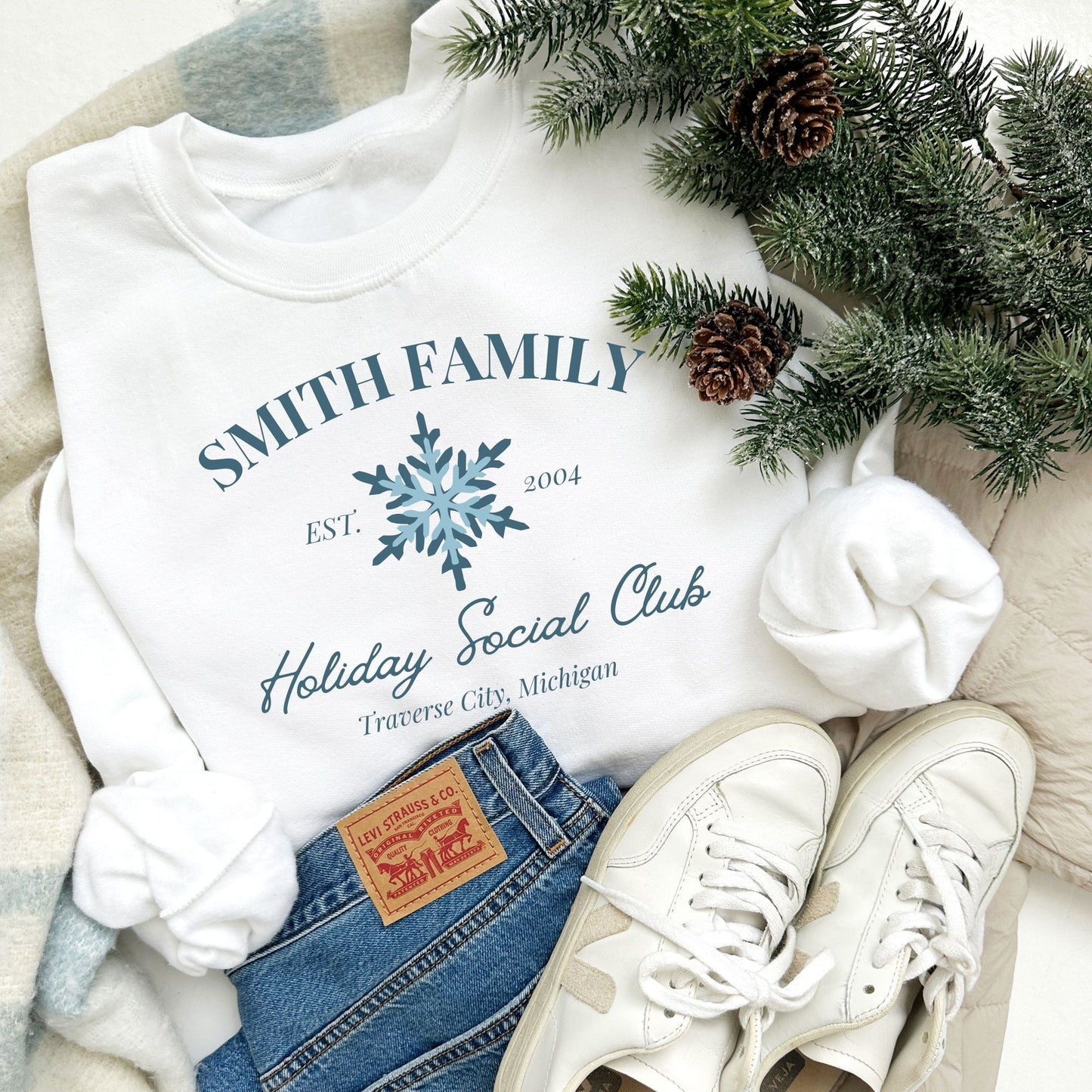 white crewneck sweatshirt with a custom print featuring a family name, Est date, snowflake, holiday social club, and location in blue