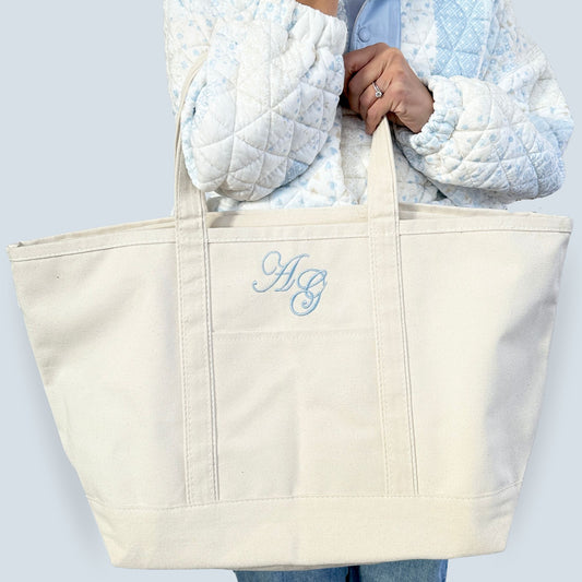 Beige tote bag with blue monogram held by a person wearing a light blue jacket.