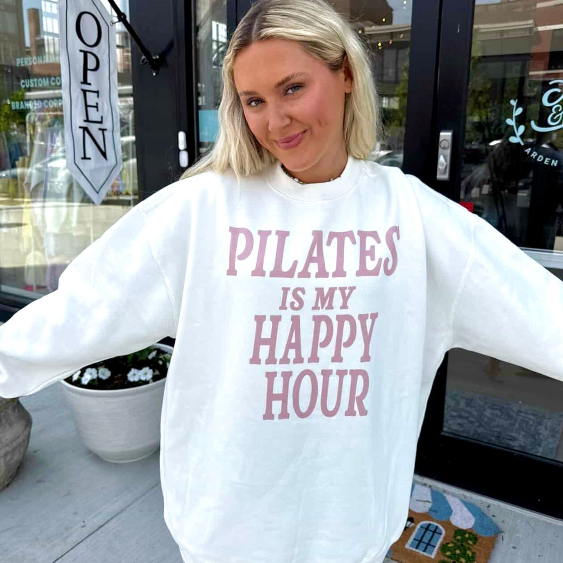 Woman wearing a white sweatshirt with 'Pilates is my happy hour' text, standing outside a building.