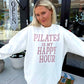Woman wearing a white sweatshirt with 'Pilates is my happy hour' text, standing outside a building.