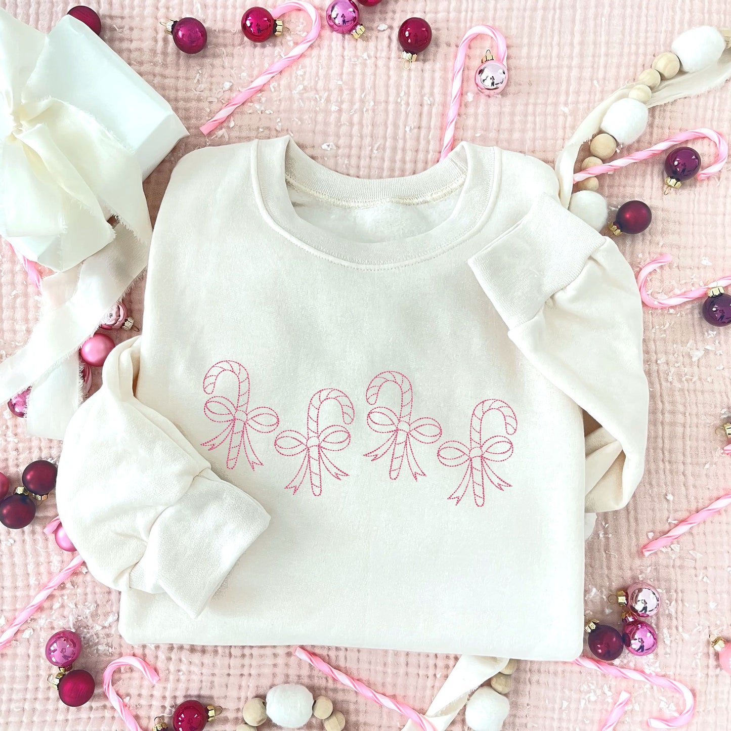 flat lay of a sweet cream crewneck sweatshirt with4 staggered outlined candy canes with bows across the chest in pink thread.