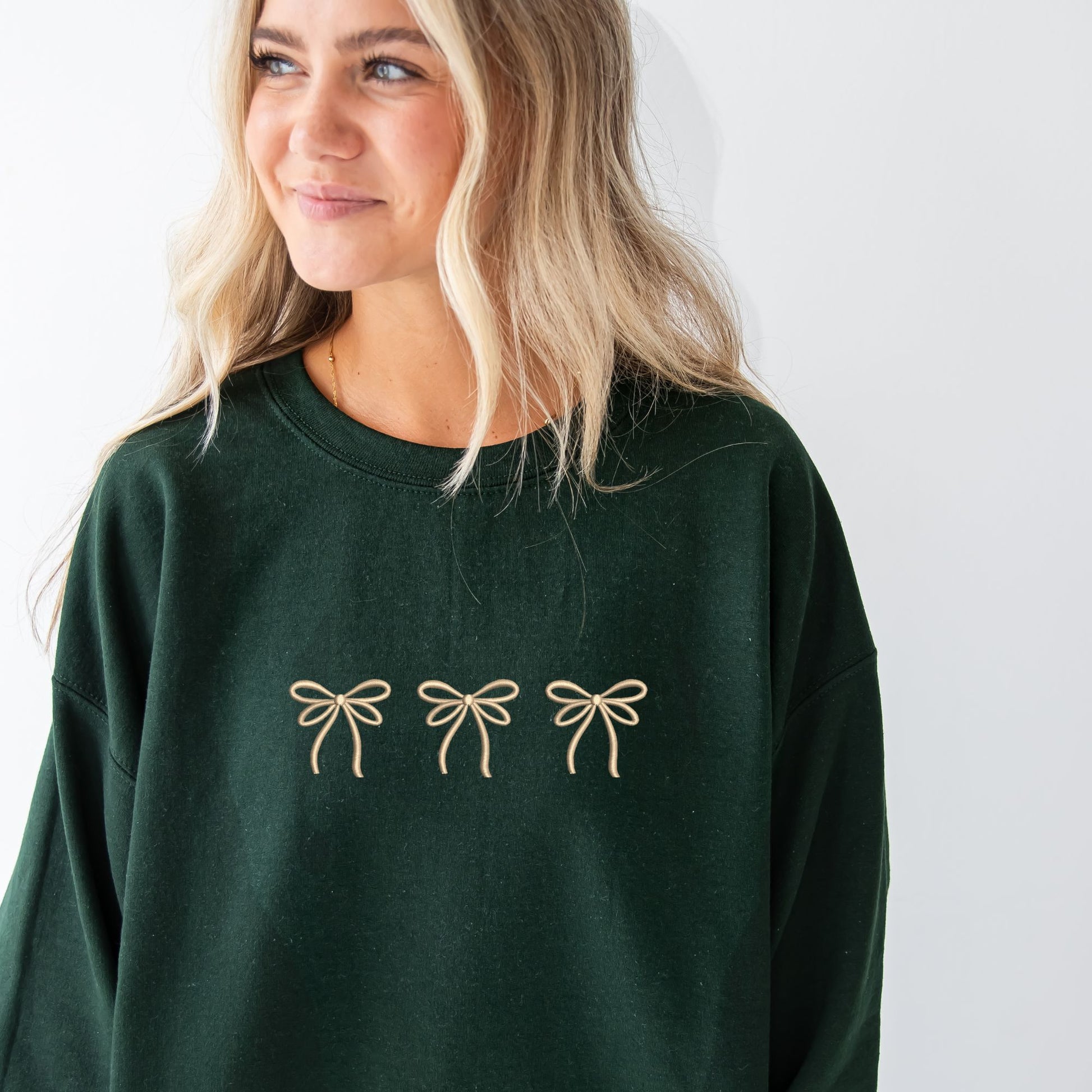 woman wearing a forest crewneck sweatshirt with embroidered three bows cross the chest in camel thread. 