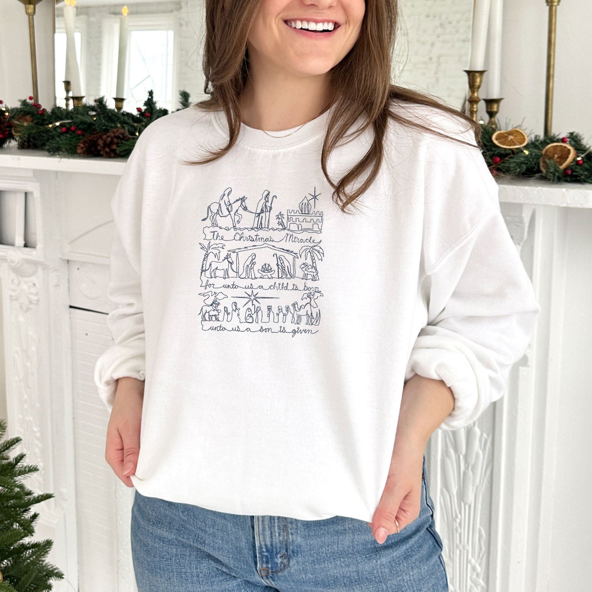 young woman wearing a white crewneck sweatshirt with embroidered stitched drawing of the Christmas nativity in navy thread.