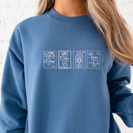 a girl standing in front of a white brick wall wearing an indigo sweatshirt with 4 embroidered Italian inspired stamps across the chest in white thread. The stamps feature lemons, a lobster, a beach scene, and seashells.