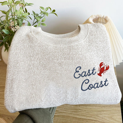 folded oatmeal heather cozy knit fleece crewneck sweatshirt with East Coast embroidered in a script font paired with a lobster on the left chest.