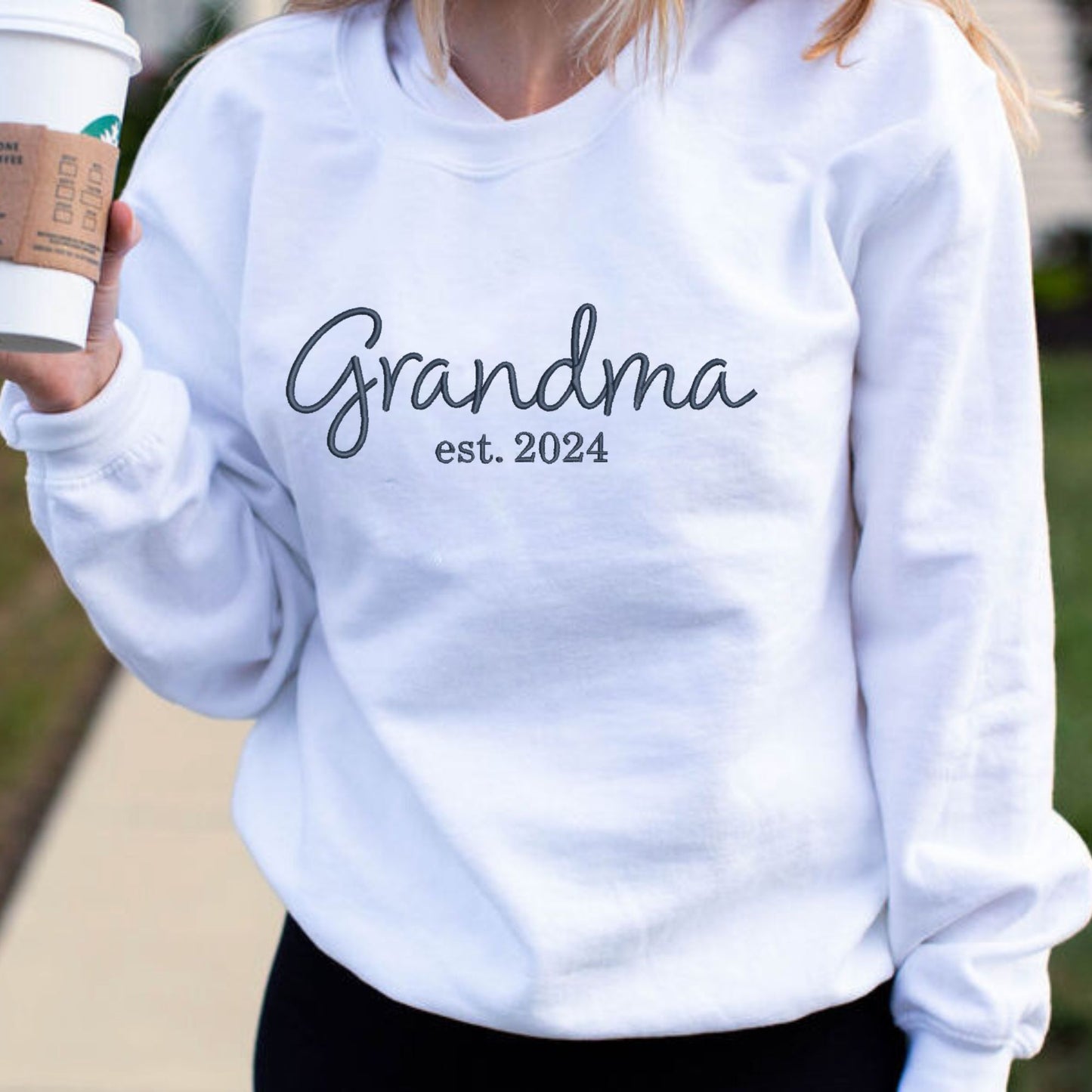 close up of a woman wearing an white crewneck sweatshirt with grandma est 2024 embroidered across the chest in navy thread