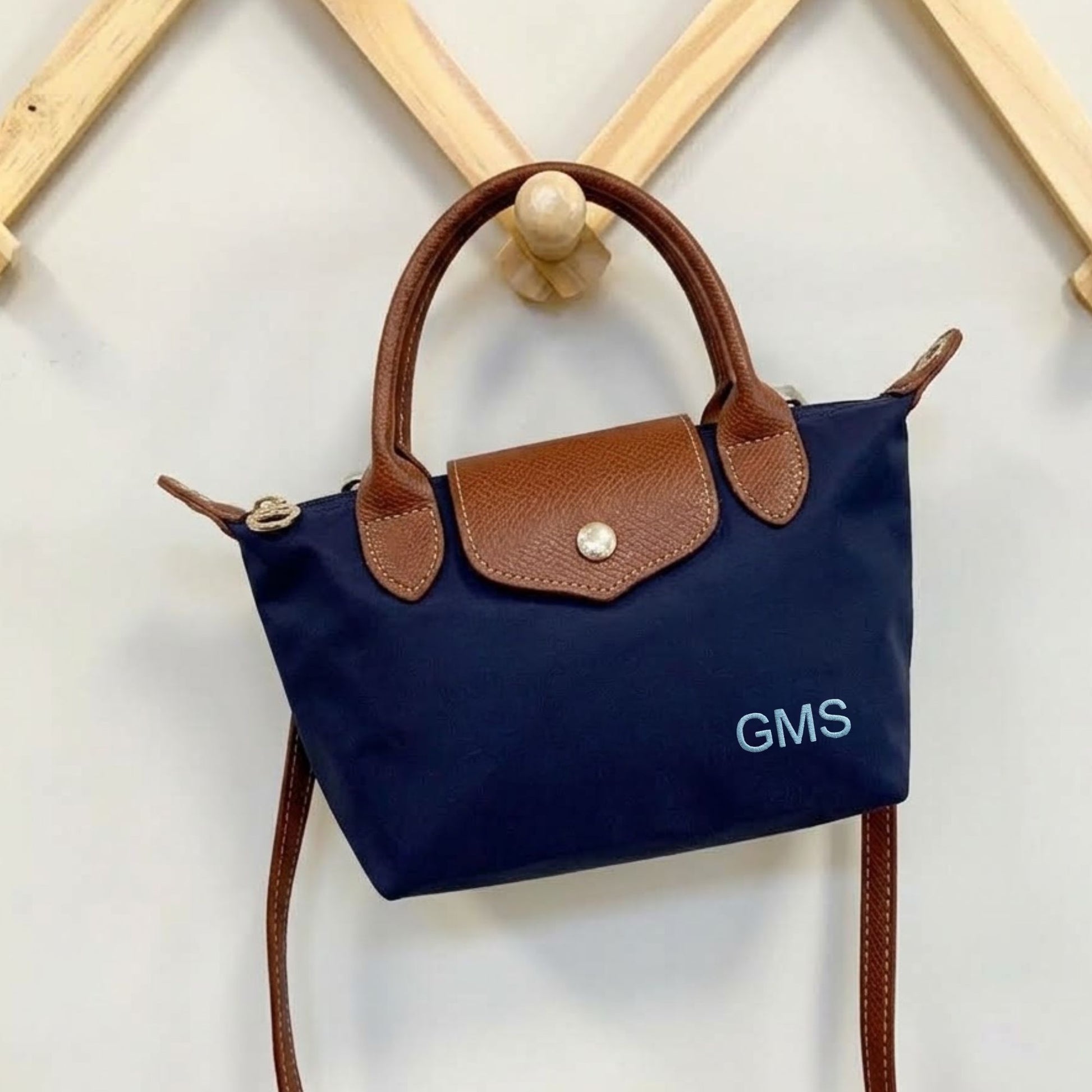 A navy blue nylon handbag with brown leather handles and flap, featuring the powder blue embroidered letters 'GMS' on the front, hanging from a wooden peg on a light wall.
