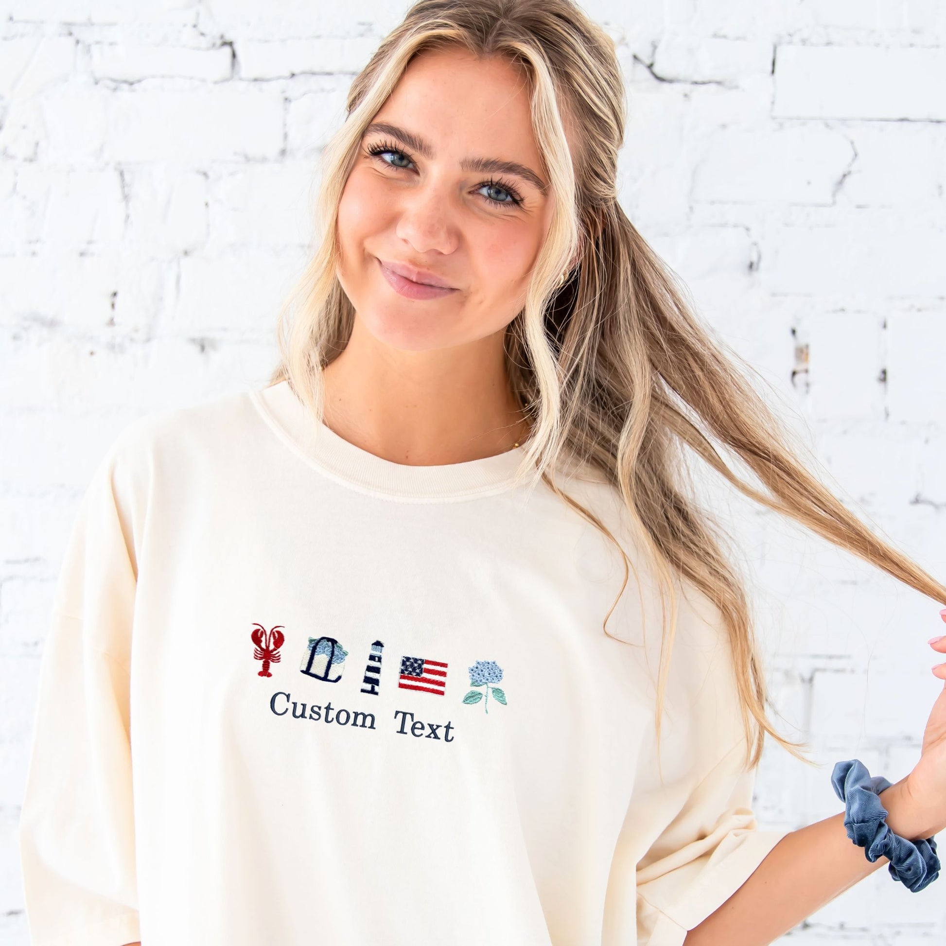Woman wearing a cream-colored sweatshirt with embroidered designs and text against a white brick wall.