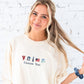 Woman wearing a cream-colored sweatshirt with embroidered designs and text against a white brick wall.
