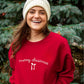 Woman wearing a red 'merry christmas' sweatshirt in front of Christmas trees.