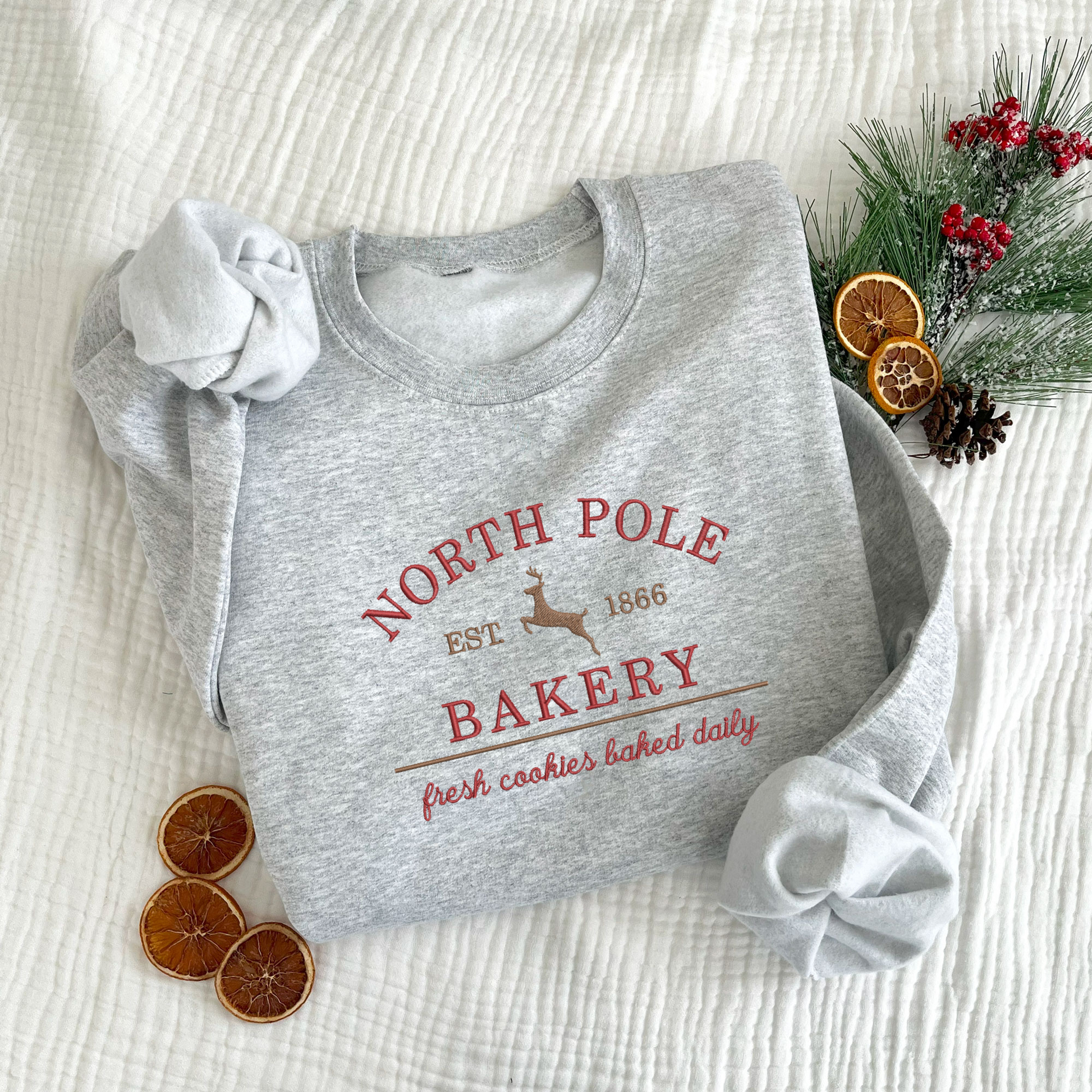 christmas styled flat lay of an ash crewneck sweatshirt with embroidered north pole bakery design in red and brown threads.