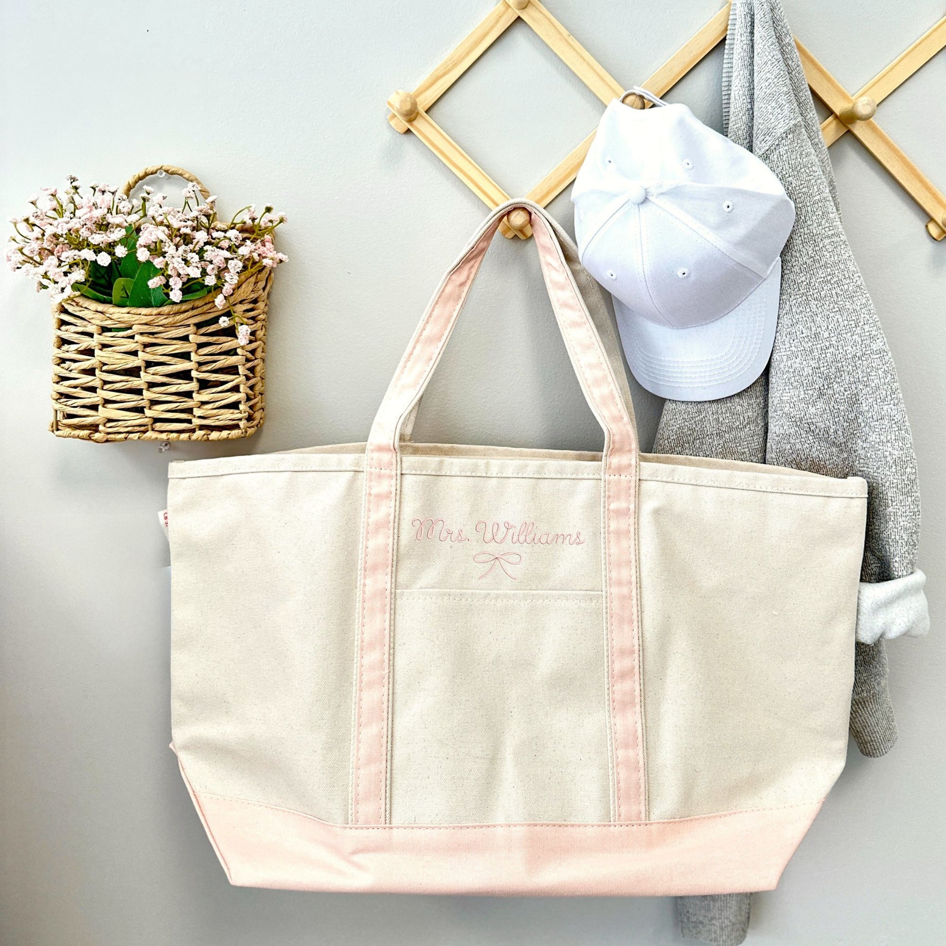 Canvas tote bag with a powder pink trim and custom name embroidery hanging on a natural wood hook next to a white baseball hat and a small wicker basket containing pale pink flowers