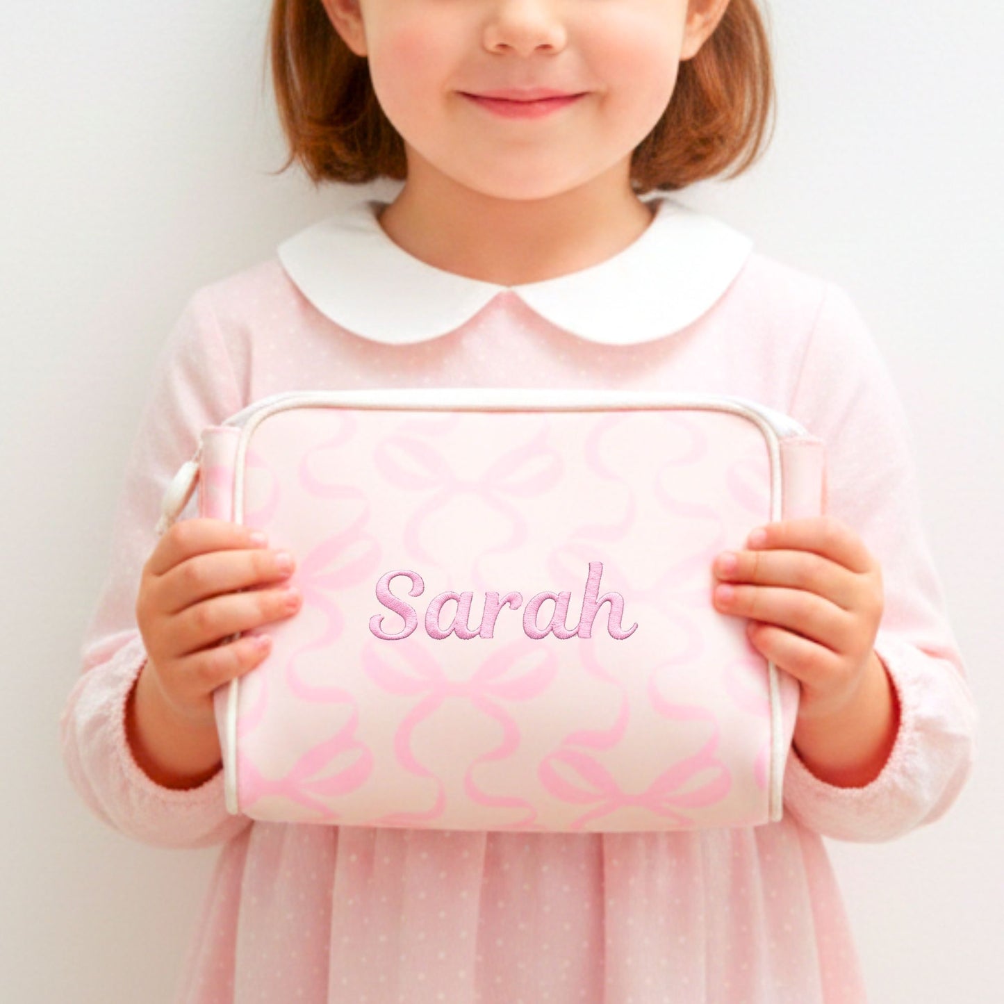 Child holding a pink bow pouch with 'Sarah' embroidered on it against a white background
