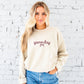 A blonde woman wearing a cream-colored crewneck sweatshirt with 'gameday' embroidered in maroon thread and a cursive font across the chest, paired with blue jeans, standing against a white brick wall.