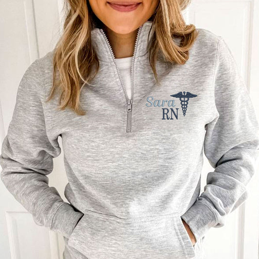 woman wearing a light heather grey quarter zip with pockets featuring a custom name, credentials, and caduceus embroidered design in shades of blue on the left chest