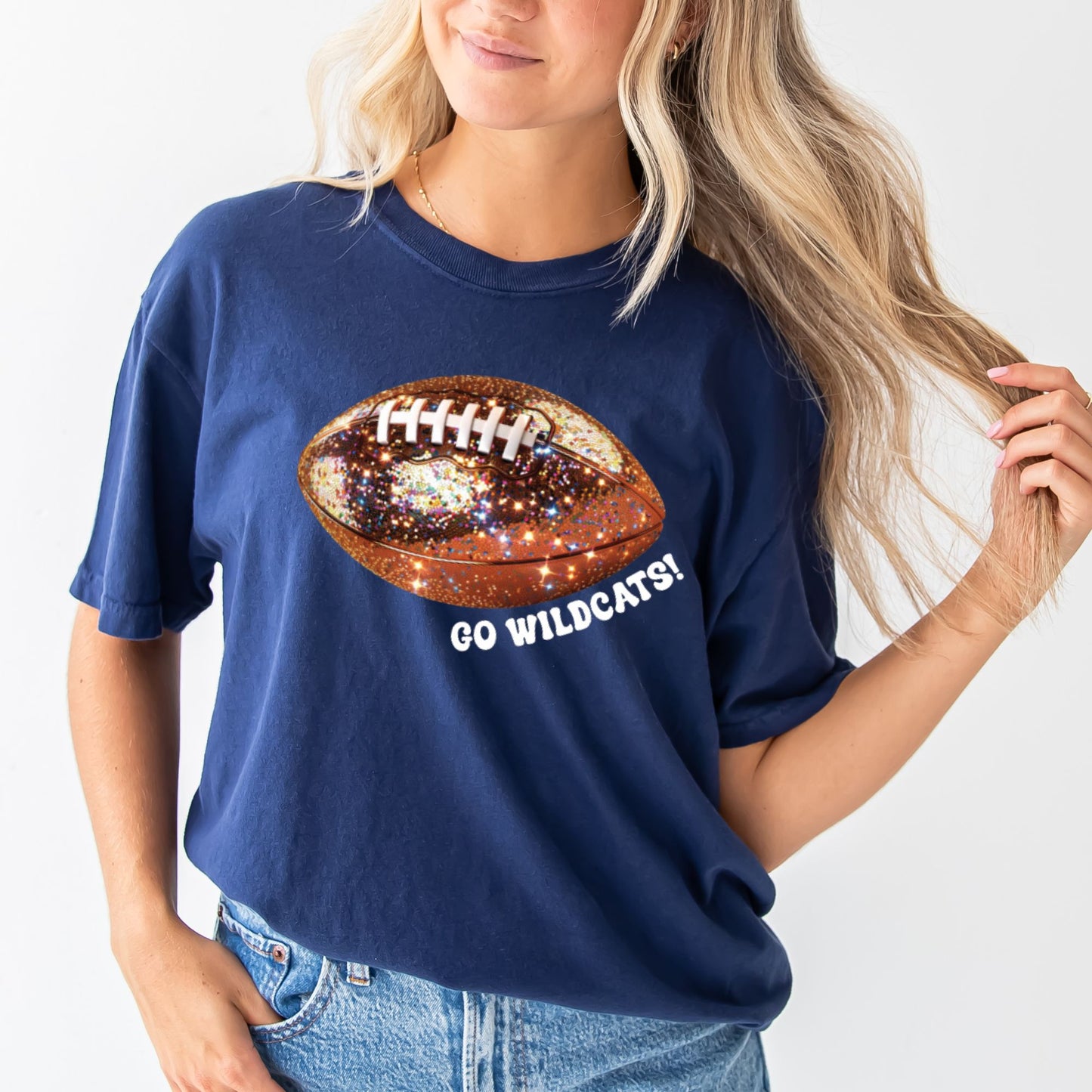 young woman wearing a navy comfort colors tee with large glitter football print and go wildcats text curved under the football in white ink