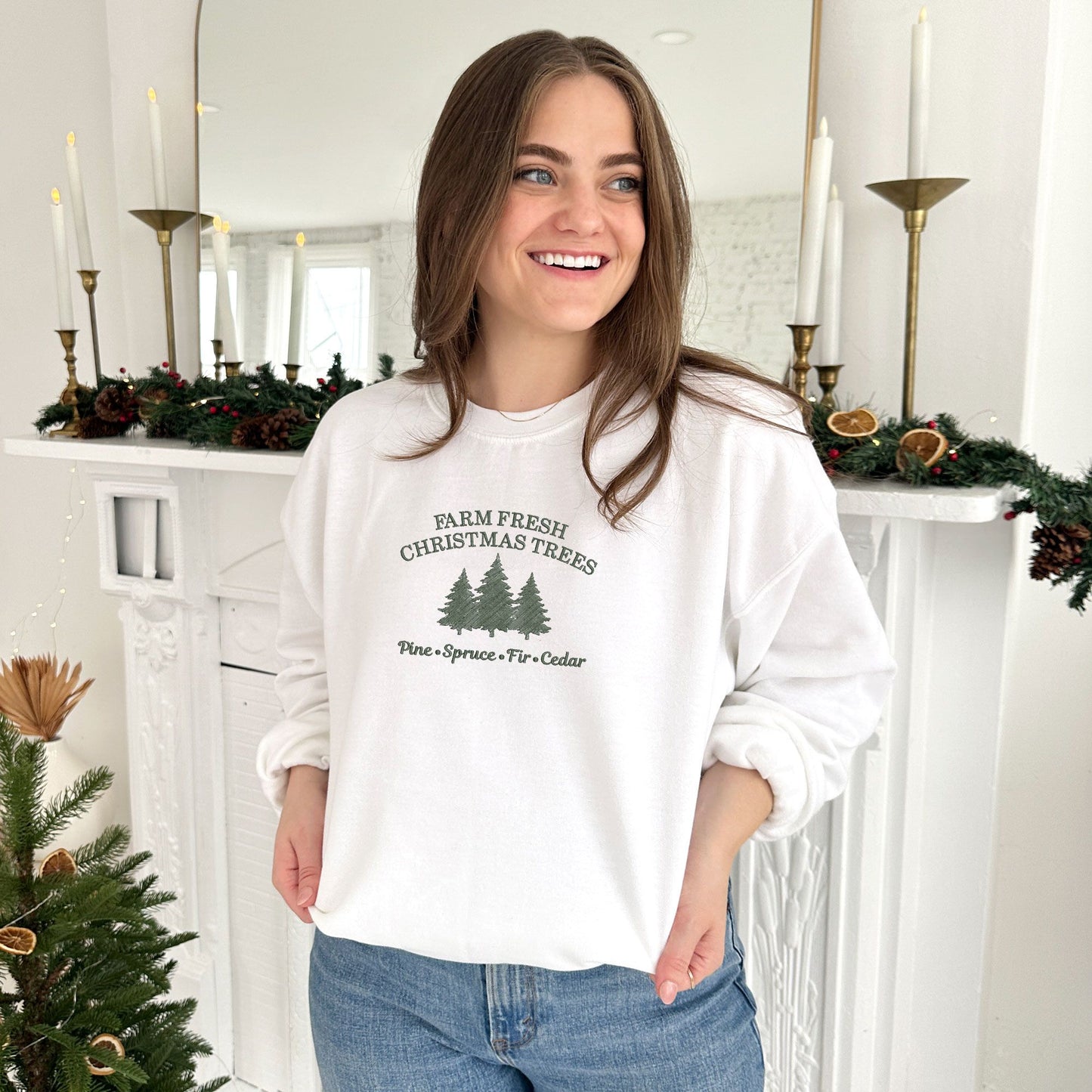 young woman wearing a white crewneck sweatshirt with embroidered farm fresh Christmas trees design with Christmas tree embroidery across the chest in ivy green thread